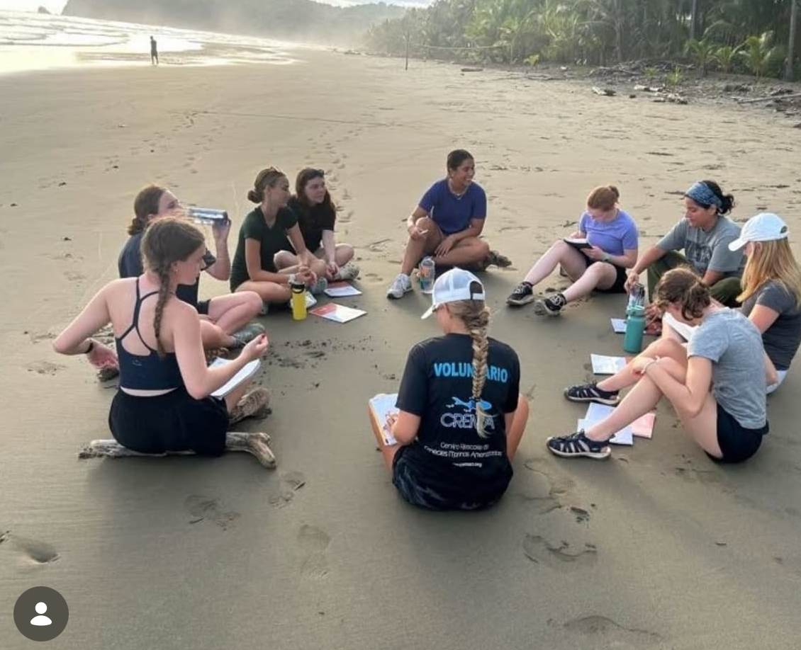 Students sitting in a circle on the sand with field notebooks, alongside a CREMA team member