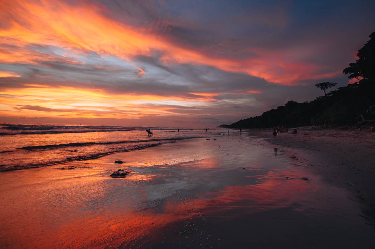 A surfer walking into the Pacific under a vivid orange and violet sunset reflected on wet sand