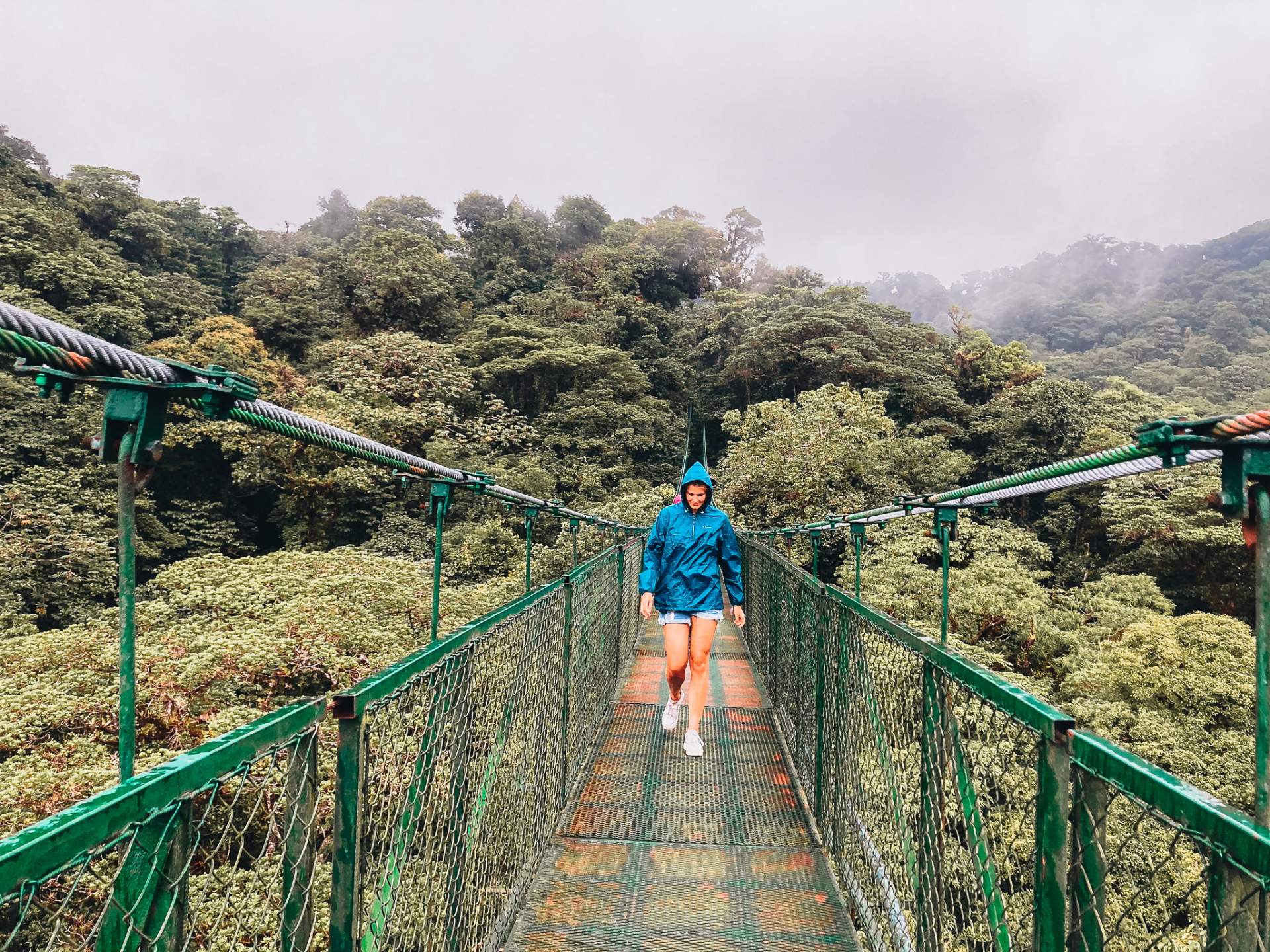 A traveler in a blue rain jacket crossing a green suspension bridge above misty cloud forest canopy