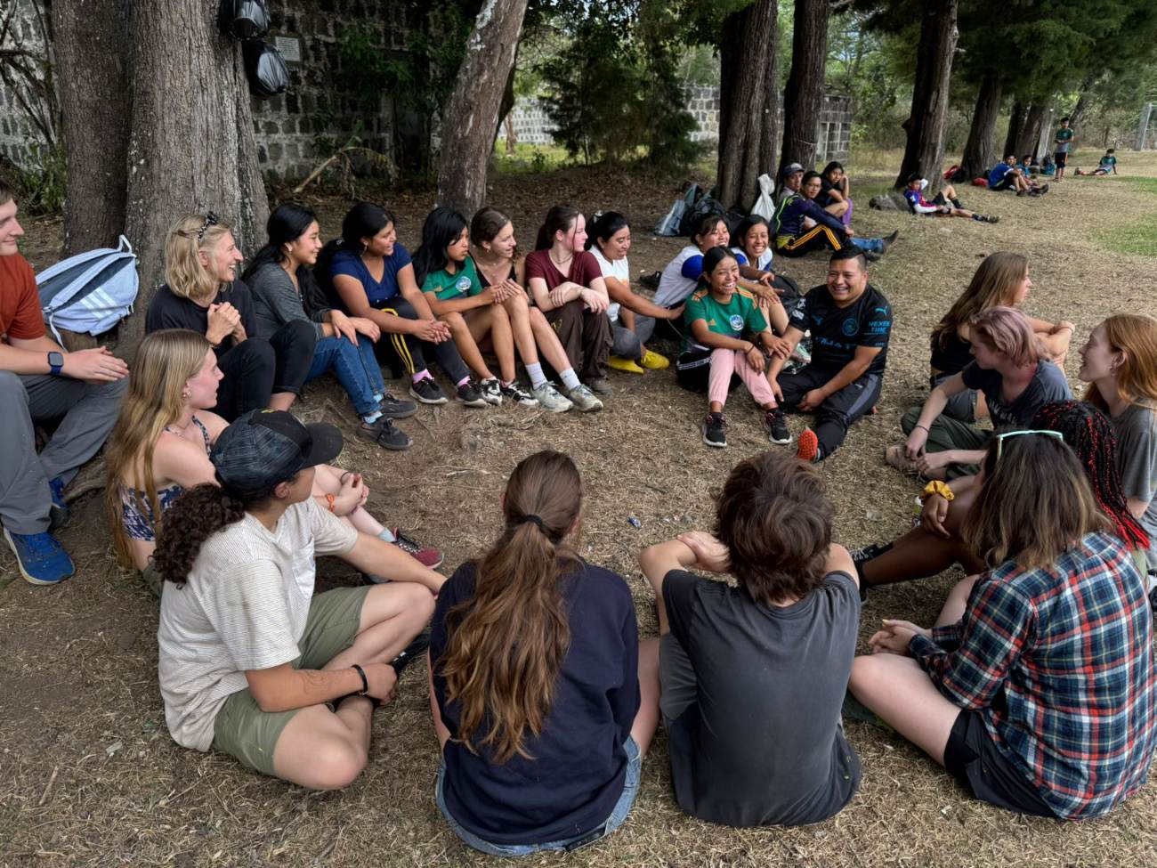 Students and local young people sitting together in a wide circle under shade trees, laughing