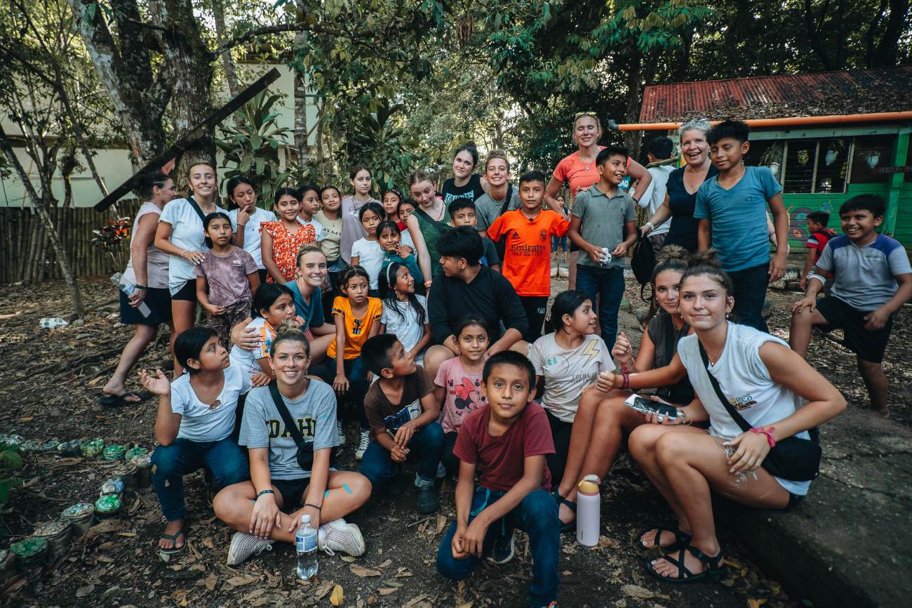 A large group of students and local children gathered together in a shaded schoolyard