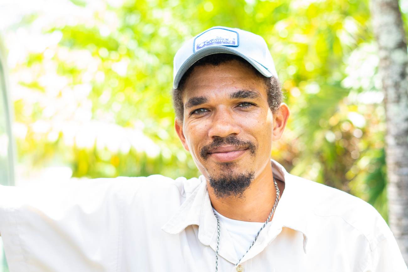 A smiling local guide in a light cap and field shirt, with bright tropical foliage behind him